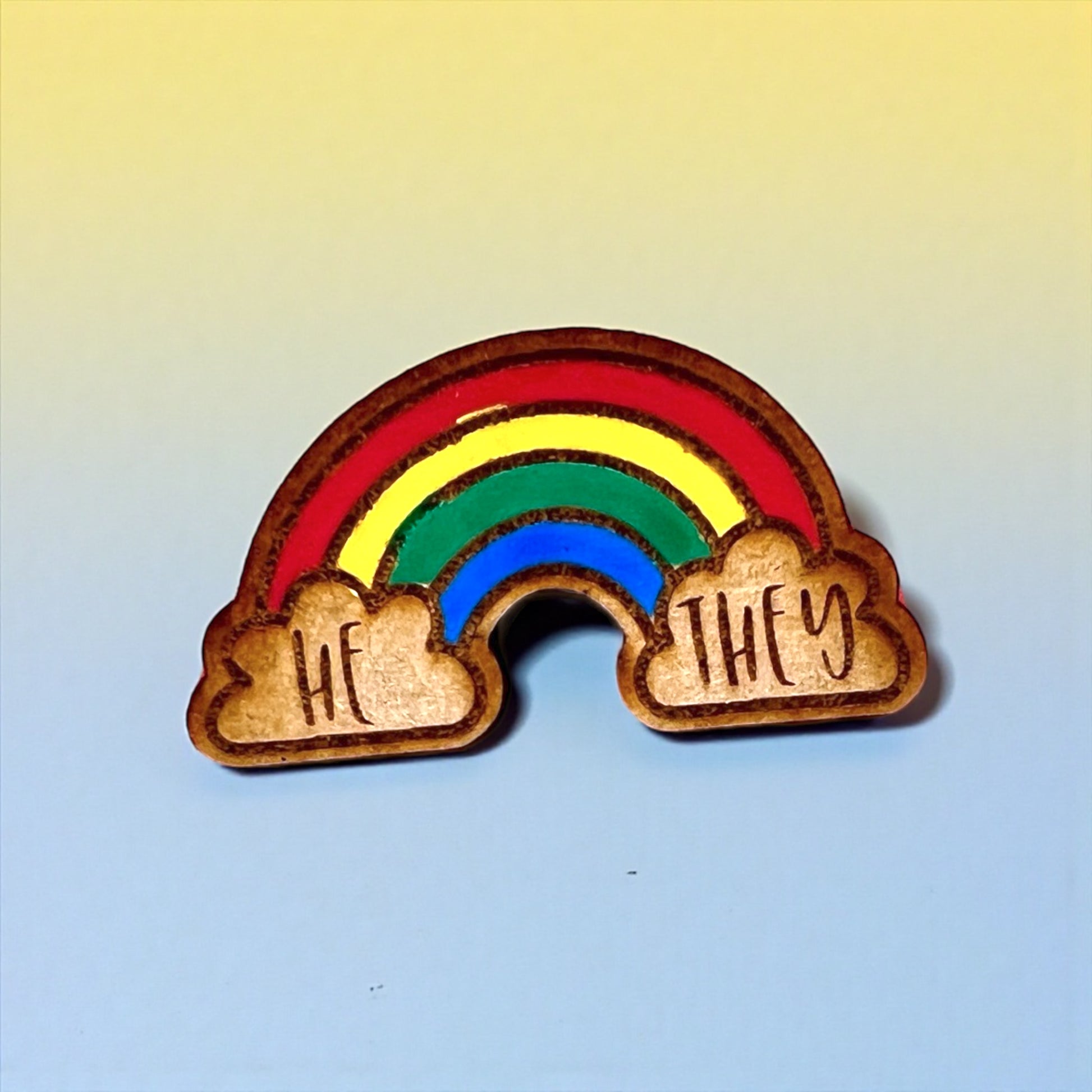 Rainbow-shaped pin with 'HE' and 'THEY' on clouds against a gradient background