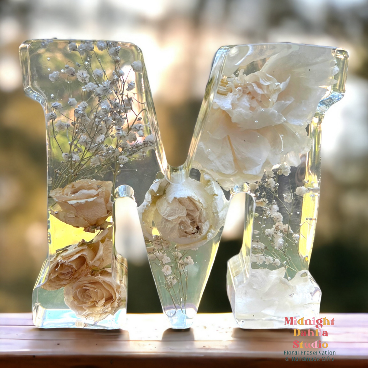 A crystal clear resin letter M with dried preserved flowers , all white , including roses , carnations , and babys breath .  A beautiful way to preserve your bridal bouquet.