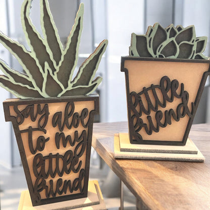 two plant pot themed decor shelf sitters in brown, black, and green, one reading say aloe to my little friend in a pretty script and the other saying little friend.