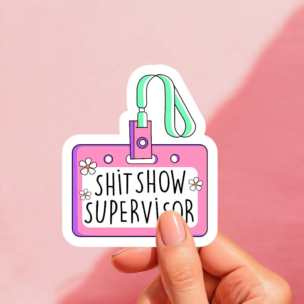 A colorful sticker with a name tag that reads 'SHIT SHOW SUPERVISOR' on a pink background, adorned with illustrations of flowers and a lanyard.