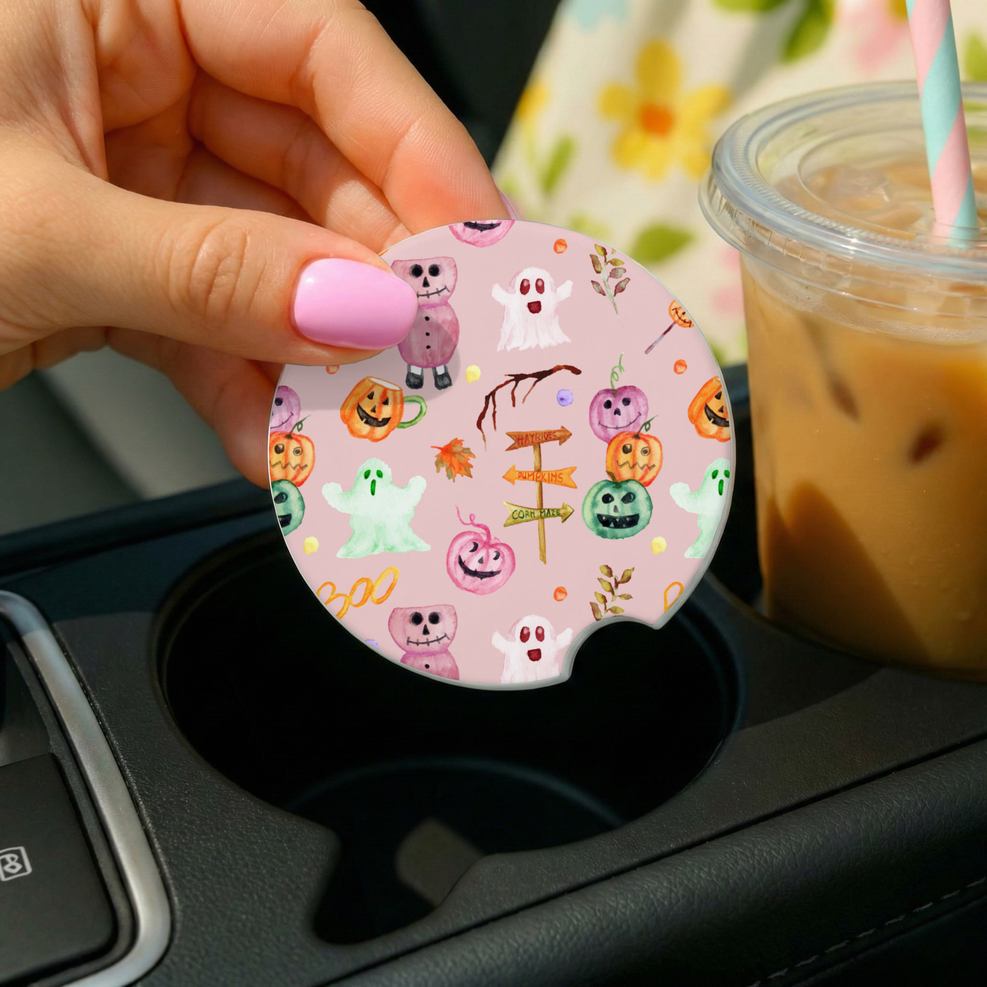 Hand holding a pink Halloween-themed car coaster over a cup holder with a drink in the background.