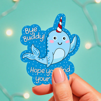 Hand holding a blue sticker with a cartoon narwhal and text on a teal background with string lights.