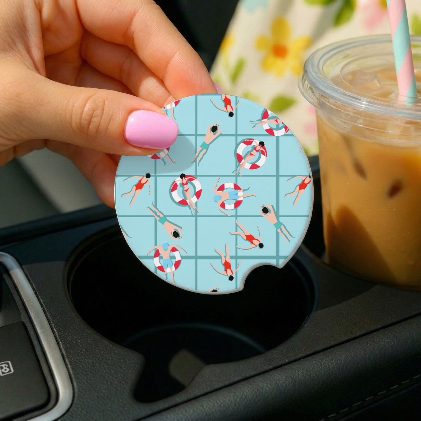 Hand holding a car coaster with a summer design over a cup holder, with a drink in the background.