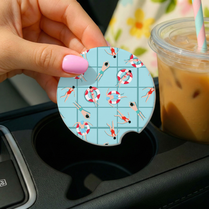 Hand holding a car coaster with a summer design over a cup holder, with a drink in the background.