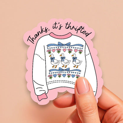 Sticker with a sweater design and text held by a hand on a beige background