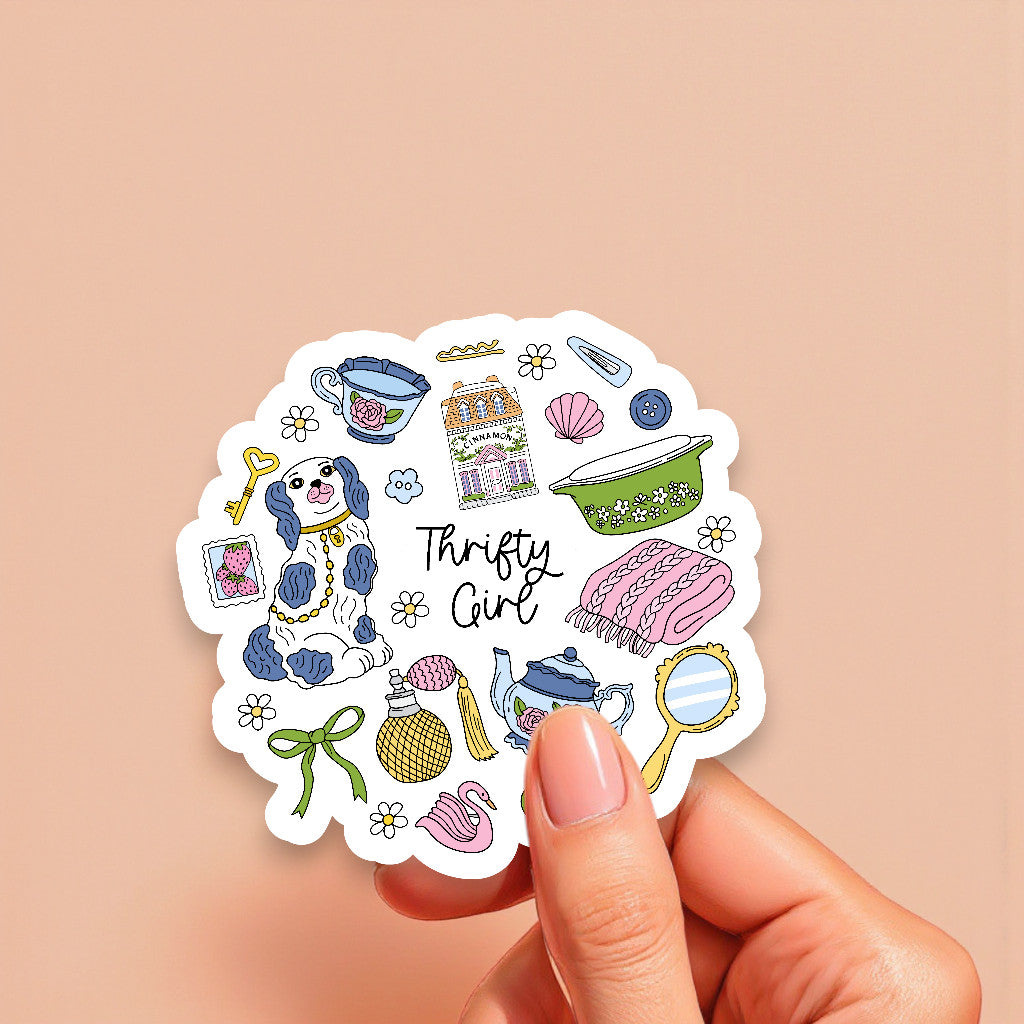 Sticker with various illustrations and the text 'Thrifty Girl' held by a hand on a beige background