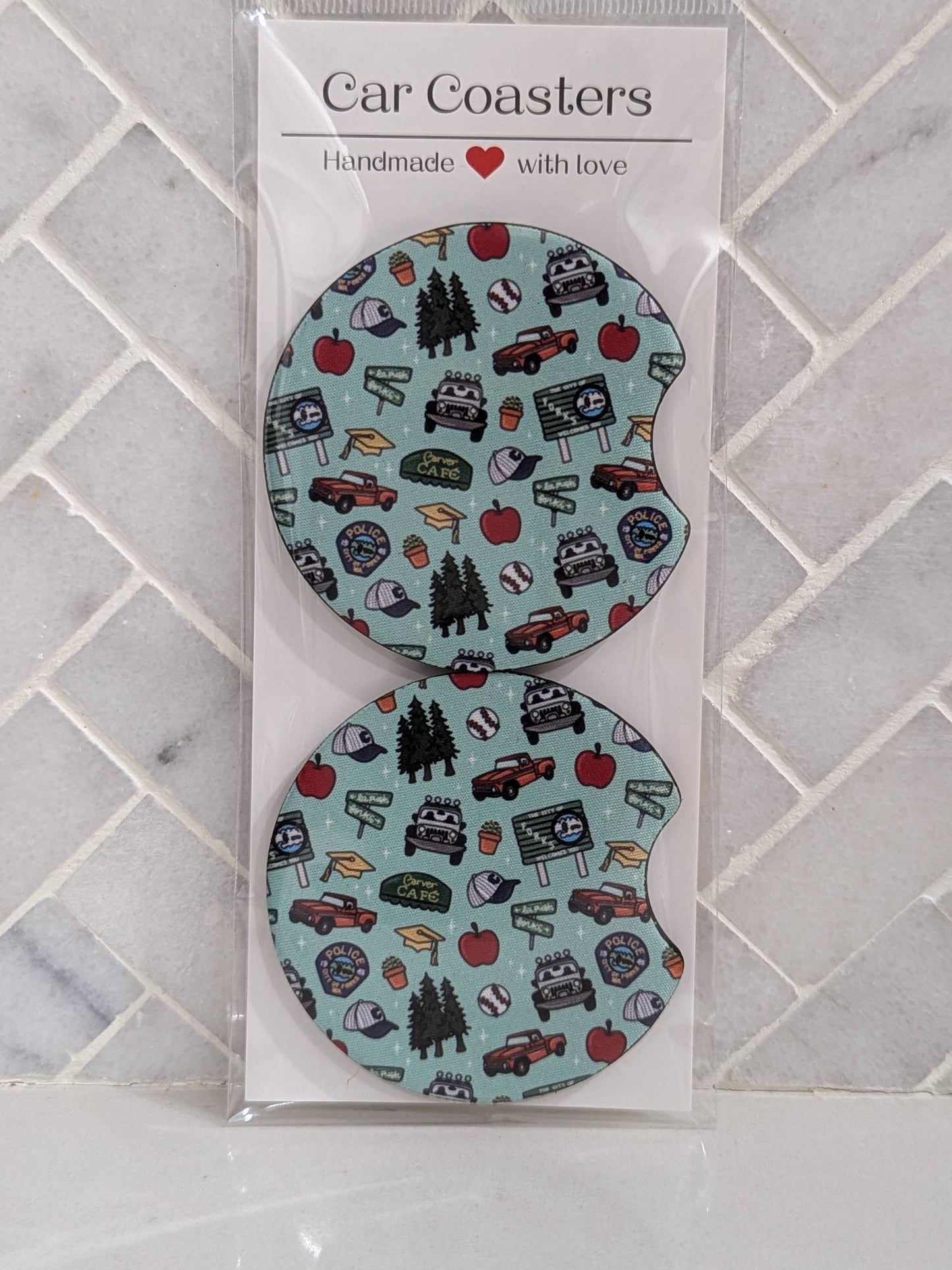 Two car coasters with a pattern of trees, cars, and other elements in a clear packaging on a tiled surface.
