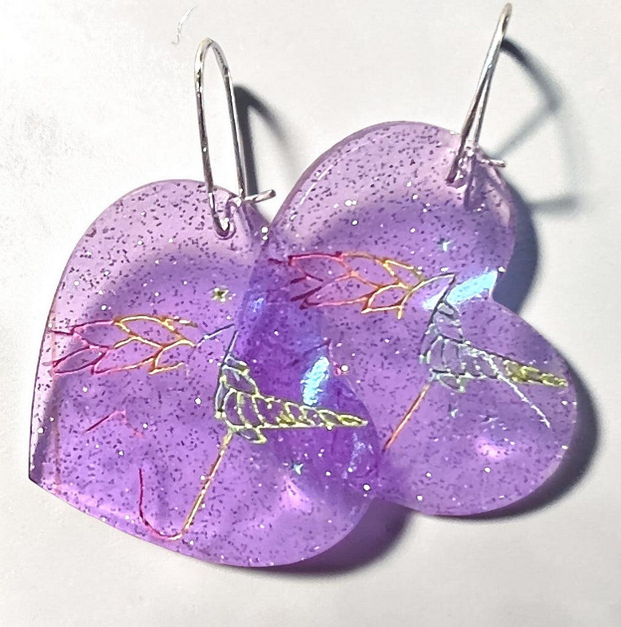 two purple glitter acrylic heart shaped dangle earrings with a rainbow unicorn head engraved.  the earrings are on silver kidney shaped hardware against a white background.