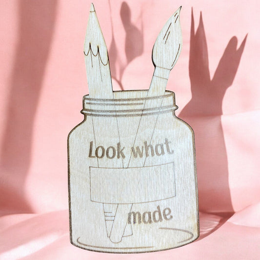 a wooden magnet that is a jar with a pencil and a paintbrush in it engraved with look what blank made to add the child's name.