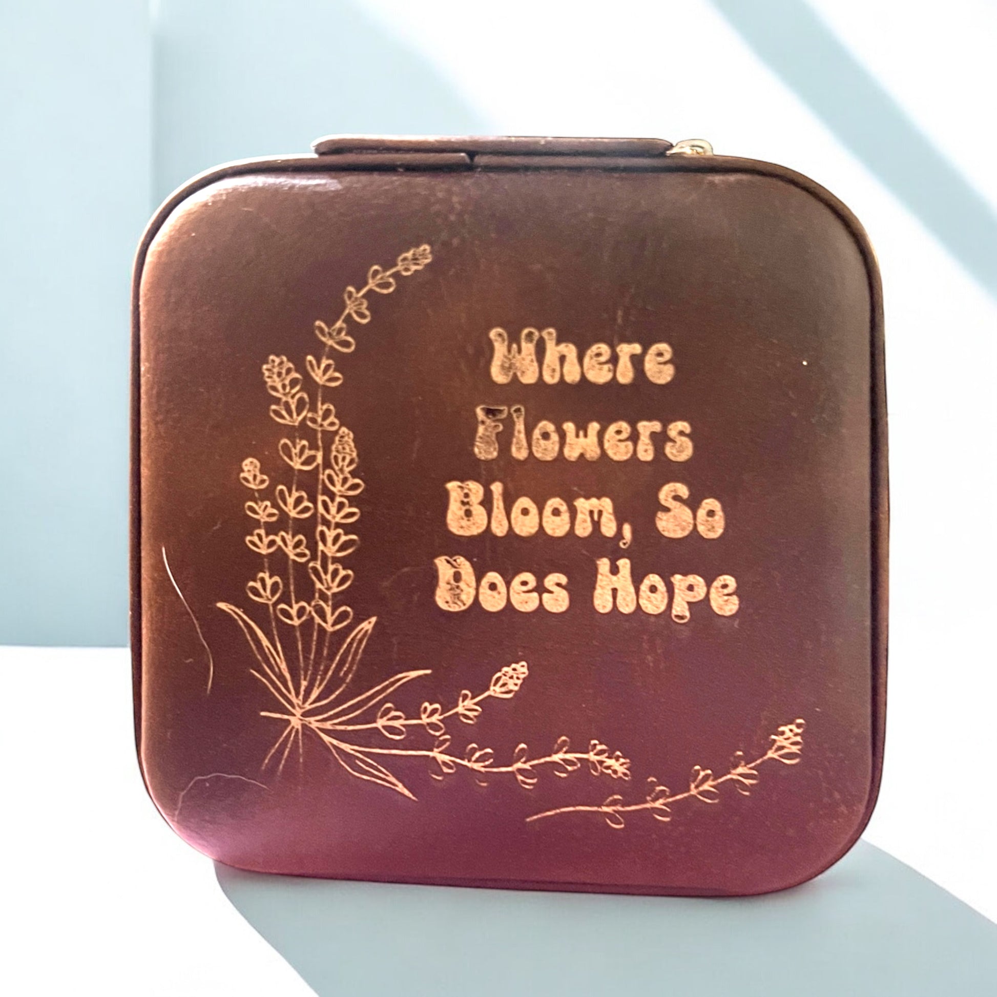 a brown jewelry box with floral accents and words engraved in gold that read 'where flowers bloom so does hope'
