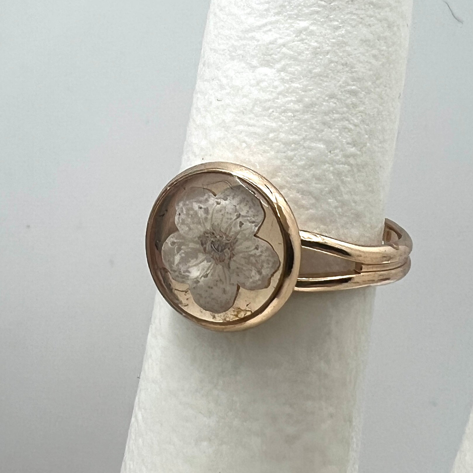 Gold ring with a floral design on a white cylindrical stand against a light gray background