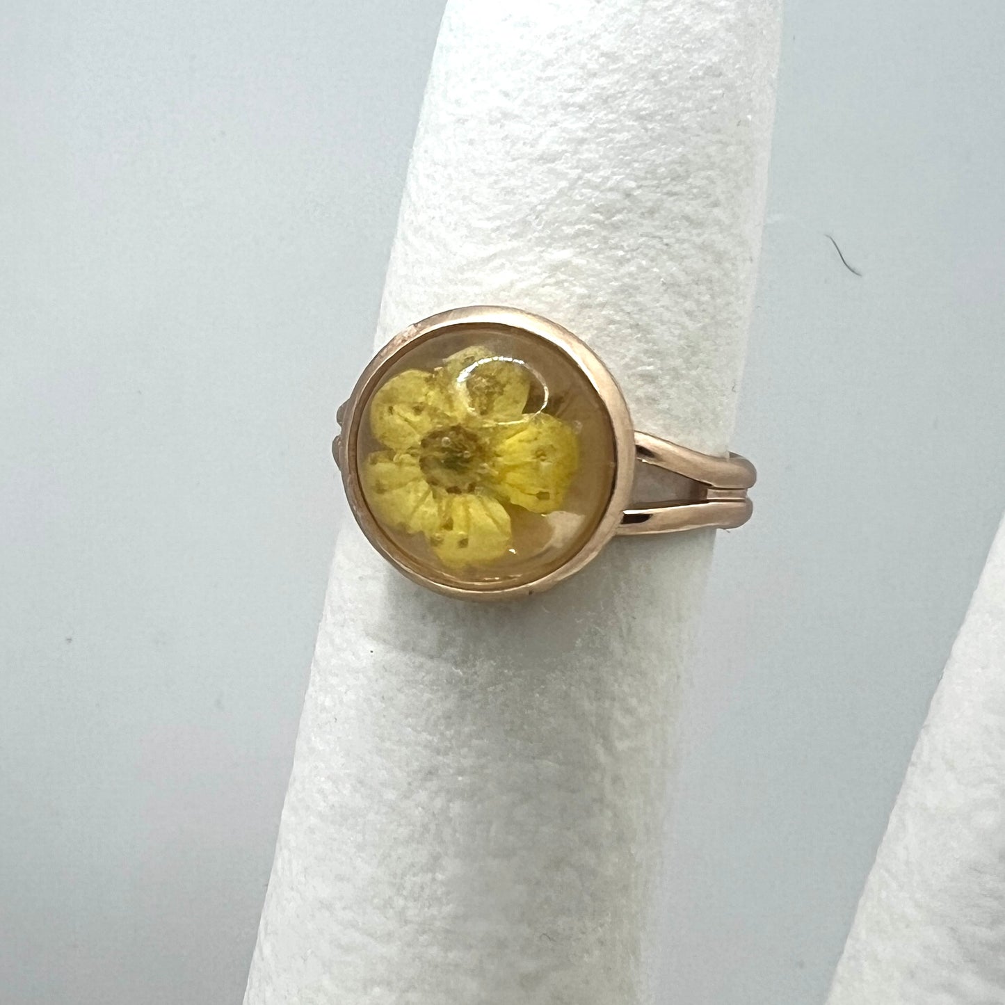 Gold ring with a yellow forget me not real pressed flower on a white stand against a light gray background