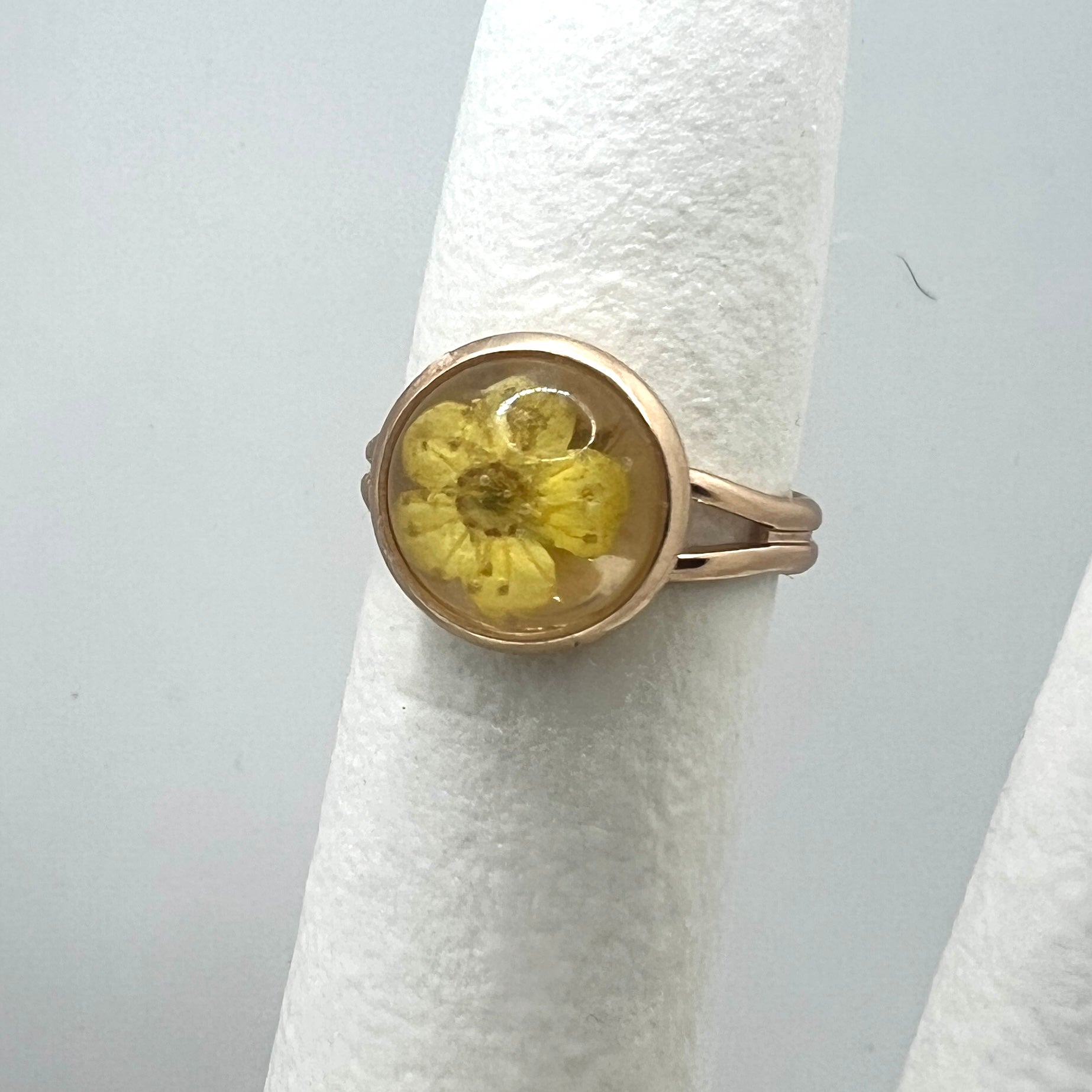 Gold ring with a yellow forget me not real pressed flower on a white stand against a light gray background