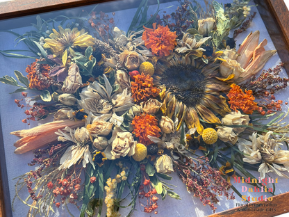 a wooden framed shadow box filled with many flowers, including yellow brown sunflowers, orange lilies, orange marigolds, white roses, white dahlias, yellow billy balls, yellow solidago, yellow dahlias, mini spray roses, green leaves, and more.