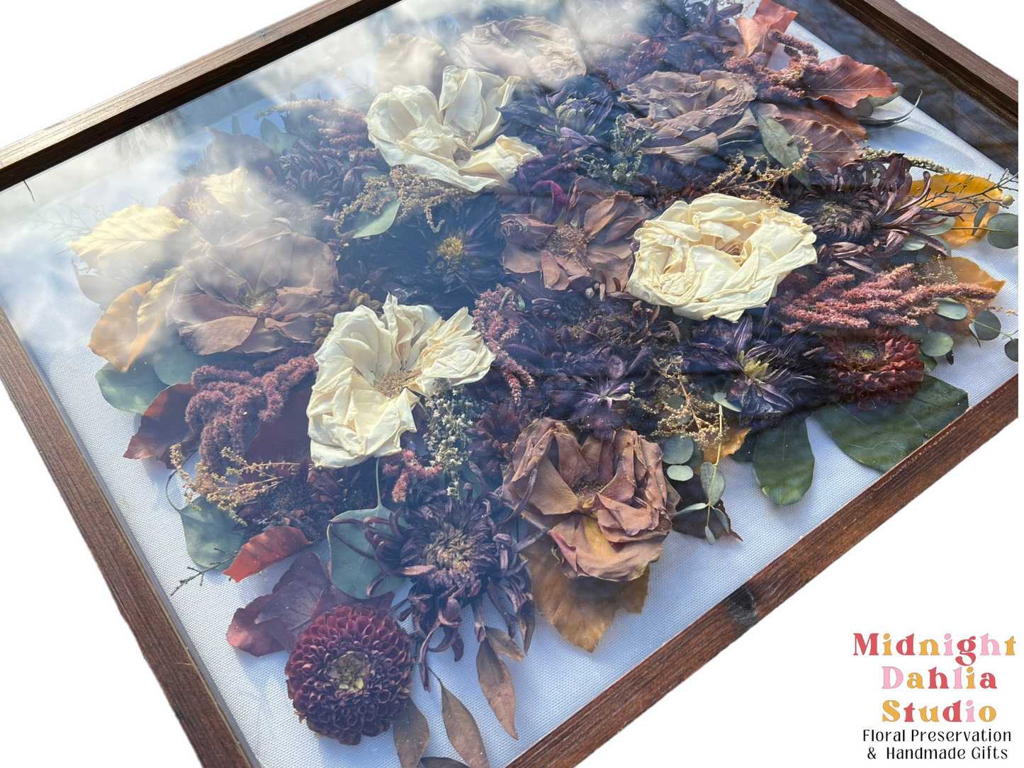 a wooden framed shadow box filled with many flowers, including white roses, taupe brown roses, burgundy red zinnia, burgundy dahlia, red amaranthus, fall leaves, and green leaves.