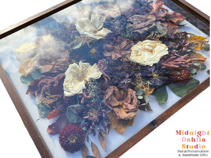 a wooden framed shadow box filled with many flowers, including white roses, taupe brown roses, burgundy red zinnia, burgundy dahlia, red amaranthus, fall leaves, and green leaves.
