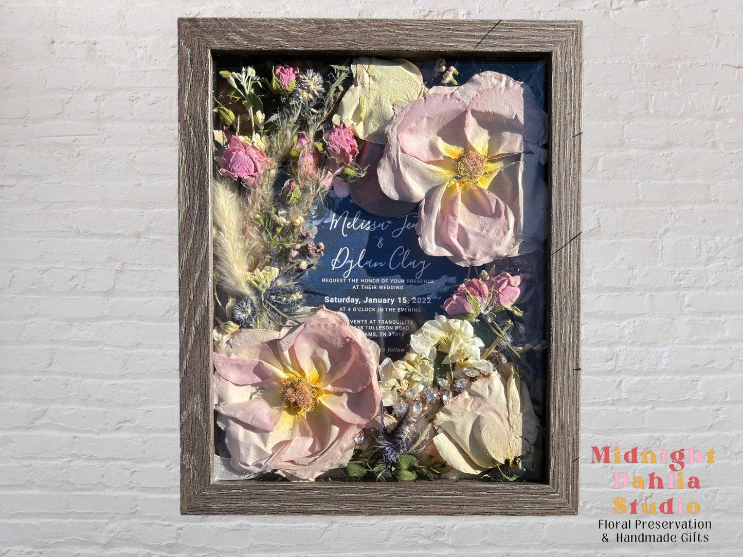 a wooden framed shadow box filled with many flowers, including pink roses, pink mini spray roses, jewels, white stock, green leaves, and an invitation.