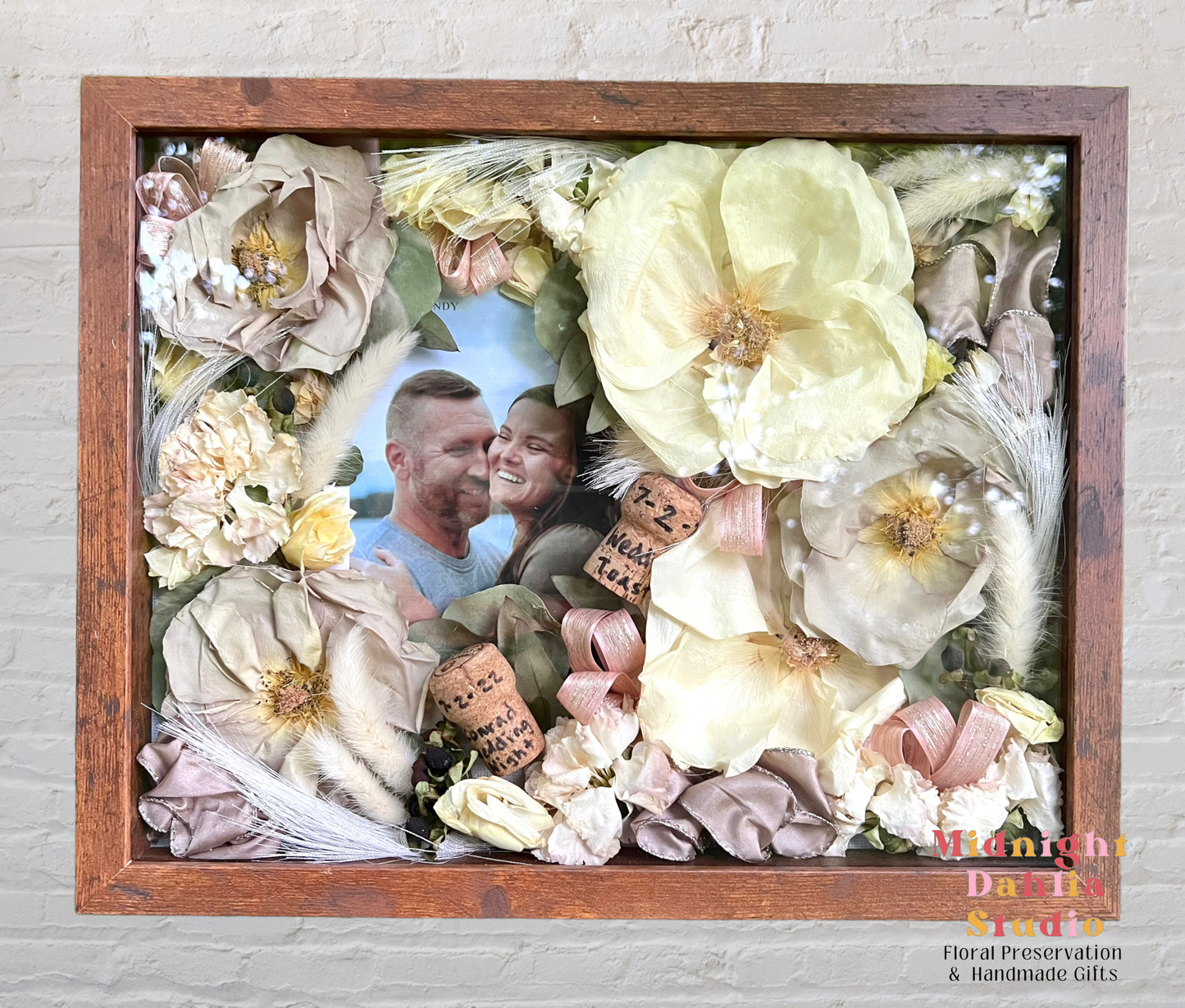 a wooden framed shadow box filled with many flowers, including white roses, pale pink roses, ribbon, corks, and a picture of a happy couple.