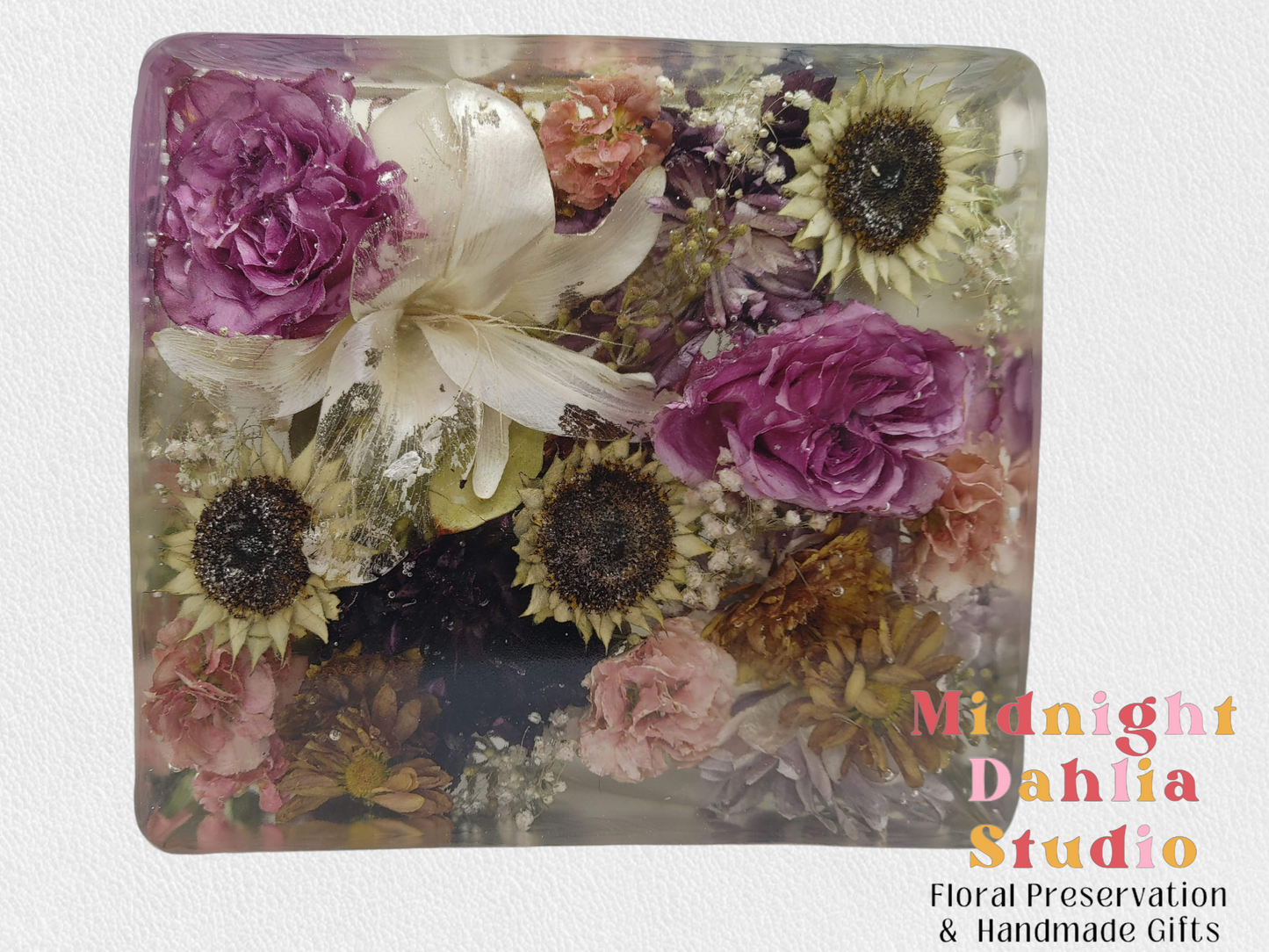 A crystal clear resin block filled with pink roses , white lilies , sunflower centers , babys breath , and other flowers .  A beautiful way to preserve a bridal bouquet.