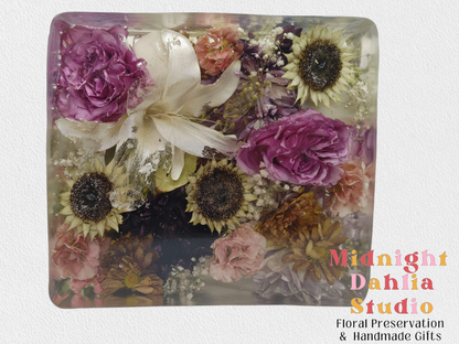A crystal clear resin block filled with pink roses , white lilies , sunflower centers , babys breath , and other flowers .  A beautiful way to preserve a bridal bouquet.